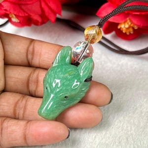 May include: A green carved stone pendant in the shape of a wolf's head. The pendant is strung on a black cord with clear and yellow beads. The background features red flowers.