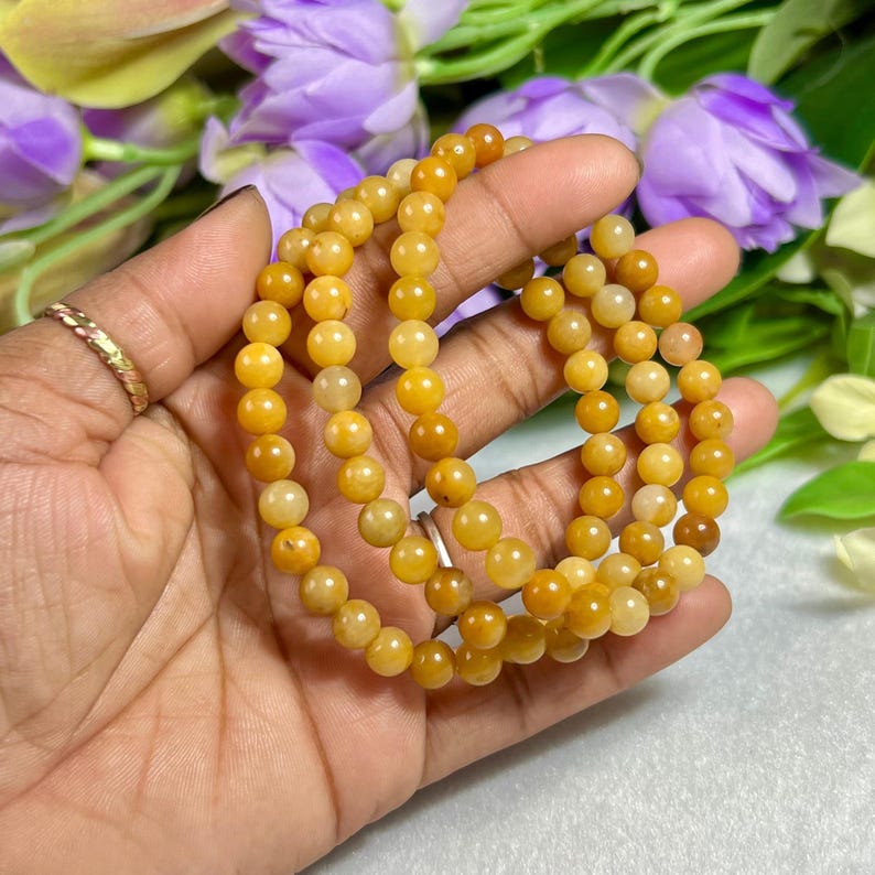 May include: A close-up of three strands of yellow-orange gemstone bead bracelets. The beads are round and have a polished finish. The bracelets are held in a hand, with a gold ring visible. Purple flowers are in the background.