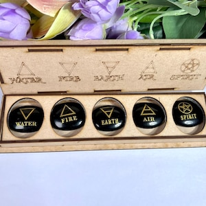 May include: A wooden box with a hinged lid, containing five black stones. Each stone has a gold symbol representing the elements: Water, Fire, Earth, Air, and Spirit. The box and stones are displayed against a backdrop of purple flowers.