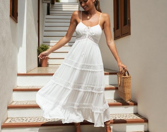 May include: A white maxi dress featuring thin spaghetti straps, a lace-up bodice detail, and horizontal lace trim inserts across the tiered skirt. The dress has a flowing silhouette and is paired with a rectangular woven straw handbag.