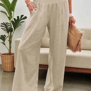 May include: Beige linen palazzo pants with a smocked waistband and wide legs. The pants have pockets and are paired with a woven clutch. The model is standing in front of a neutral-toned sofa and a potted plant.