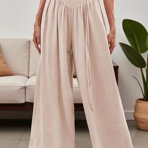 May include: Beige wide-leg pants with a high waist and a matching fabric belt. The pants have a textured fabric and a relaxed fit. The model is wearing a white crop top. The pants are suitable for casual or semi-formal wear.