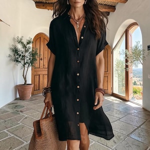 May include: A black button-down shirt dress with short sleeves and a collar. The dress features a full-length front button placket and a loose, straight silhouette. The fabric has a visible texture, and the garment is worn with a woven straw tote bag.