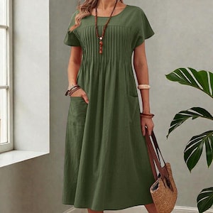 May include: Olive green midi dress with short sleeves and a round neckline. The dress features pleating on the bodice and two front pockets. A long beaded necklace and bracelets are visible. A woven tote bag is held in the hand.