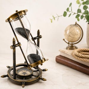 May include: An antique-style brass hourglass with black sand, mounted on a rotating frame attached to a ship's wheel base. The base features an integrated compass with a visible needle and cardinal directions, finished in a dark, weathered metal.