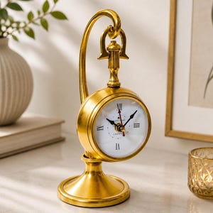 Puede incluir: Un reloj de mesa de metal dorado con esfera circular, fondo blanco y números romanos negros. El reloj cuelga de un brazo curvo unido a una base circular. La esfera presenta manecillas negras para las horas y minutos, y un segundero de color rojo.