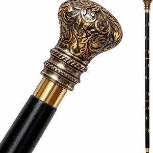 May include: A walking cane featuring a black shaft with small gold-colored diamond accents and a decorative, ornate brass-toned handle. The handle displays intricate embossed scrollwork patterns and a polished finish, connected to the shaft by a smooth gold-colored metal collar.