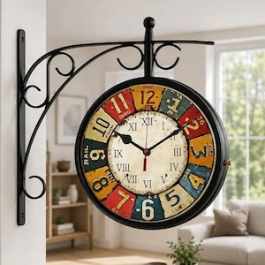 May include: Double-sided, vintage-style wall clock with a black metal frame. The clock face features a cream-colored background with black Roman numerals and colorful number blocks. The clock is mounted on a decorative black metal bracket.