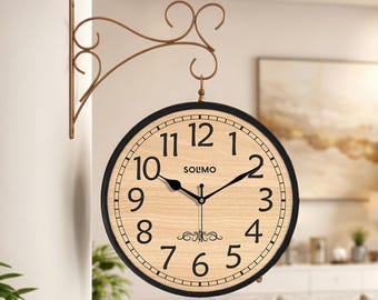 Könnte beinhalten: Eine doppelseitige Wanduhr mit einem hellen Holzmaserungszifferblatt, schwarzen Ziffern und schwarzen Zeigern. Die Uhr zeigt den Markennamen SOLMO in schwarzer Schrift und ein dekoratives Ornament über der Zahl sechs. Sie hängt an einer bronzefarbenen Halterung.