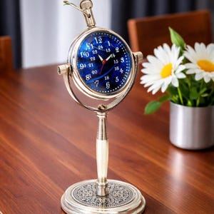 May include: A metallic desk clock featuring a circular blue face with white numerals from 1 to 12 and 13 to 24. The clock is mounted on a swivel frame atop a slender stem and a round, ornate base with intricate engraved patterns in silver tones.
