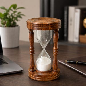 Puede incluir: Un reloj de arena con un marco de madera pulida de color marrón oscuro, con pilares torneados y bases circulares en la parte superior e inferior. El bulbo de vidrio transparente contiene arena blanca fina, acumulada en la cámara inferior.