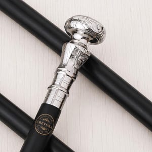 May include: A black walking cane featuring a polished silver-toned handle with intricate engraved detailing. The handle has a rounded top and a textured neck. A circular black and gold logo with the text CREVORA is printed on the shaft.