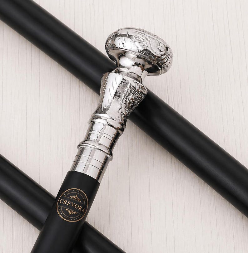 May include: A black walking cane featuring a polished silver-toned handle with intricate engraved detailing. The handle has a rounded top and a textured neck. A circular black and gold logo with the text CREVORA is printed on the shaft.