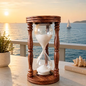 May include: A wooden hourglass with a dark brown, polished finish and turned pillars. The clear glass bulbs contain white sand, with the top bulb partially filled and the bottom bulb holding a small mound of sand, centered on a circular base.