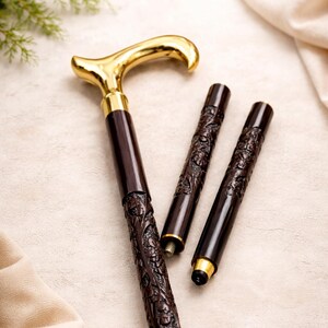 May include: A collapsible walking cane featuring a polished gold-tone derby handle and a dark brown shaft with intricate floral carvings. The cane is shown in three segments, with threaded metal connectors visible for assembly and a black rubber tip.