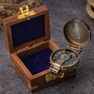 Puede incluir: Una brújula de estilo vintage con acabado de latón, abierta para revelar la esfera direccional. La brújula está alojada en una caja de madera con cierre y bisagras de latón, forrada con terciopelo azul. La caja está abierta.