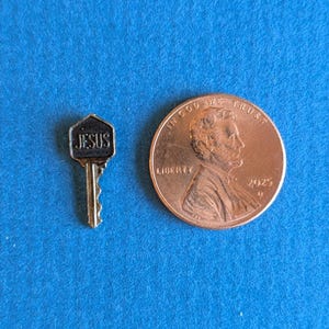 May include: A small metallic key-shaped pin with a hexagonal head featuring the word JESUS in raised letters, positioned next to a copper-colored United States one-cent coin for scale, showing the profile of Abraham Lincoln and the year 2025.