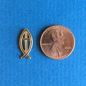 May include: A small gold-toned metal pin shaped like a fish with a cross inside, positioned next to a copper-colored 2025 United States one-cent coin for scale. The pin features a hollow design with a central vertical cross and a pointed tail.