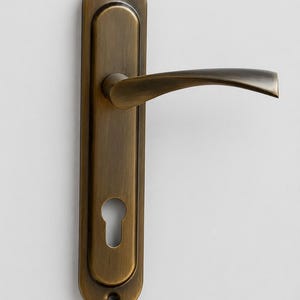 May include: A door handle set with a long, vertical rectangular backplate featuring rounded ends and a brushed antique bronze finish. The assembly includes a curved lever handle and a keyhole cutout, with visible screw holes at the top and bottom.