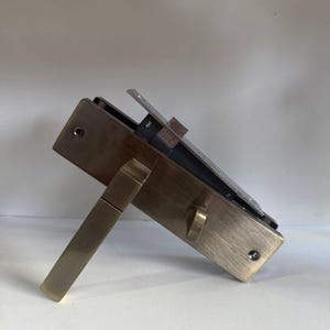 Puede incluir: Un mecanismo de cerradura de puerta rectangular con acabado en bronce cepillado. El herraje cuenta con una manija de palanca, un pequeño pomo giratorio y un pestillo metálico saliente. El conjunto incluye una caja interna y orificios para tornillos.