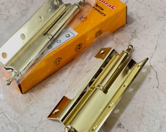 May include: Two gold-coloured door hinges, one partially in its packaging, are displayed on a marble surface. The packaging is orange with the text "Kozak" and "Door Hinges". The hinges have a decorative finial at the end.