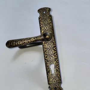 May include: Ornate bronze door handle with a floral pattern. The handle features a long, rectangular backplate with a keyhole and a curved handle. The design is detailed with a repeating floral motif, giving it a vintage aesthetic.