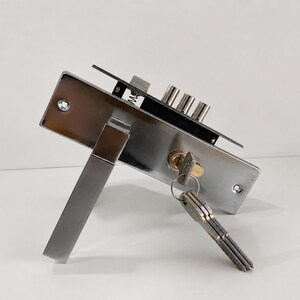 May include: A silver-colored metal door lock mechanism featuring a rectangular faceplate, a lever handle, and a brass-toned cylinder lock. Three cylindrical bolts extend from the top edge, and a metal key is inserted into the lock with a ring of keys.