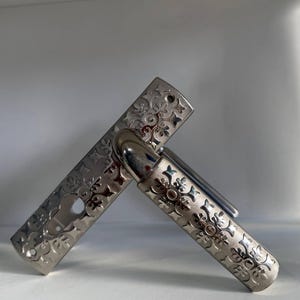 May include: A silver-toned metal door handle set featuring an elongated rectangular backplate with a keyhole cutout and a matching lever handle, both decorated with a repeating embossed pattern of stylized floral and star-like shapes.