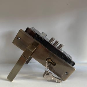 Op de afbeelding: Een messingkleurig deurslotmechanisme met een rechthoekige voorplaat, een bijpassende deurkruk en een nachtschoot met drie cilindrische stalen bouten. Een zilverkleurige metalen sleutel zit in het sleutelgat en hangt naar beneden.