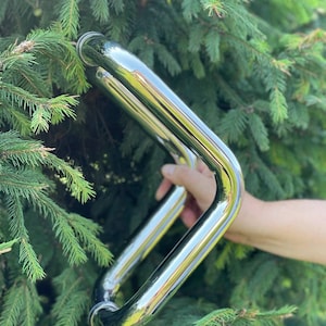 Op de afbeelding: Een glanzende, verchroomde deurklink met een gebogen, rechthoekig ontwerp. De klink wordt tegen een achtergrond van groene dennennaalden gehouden, die de omgeving weerspiegelt.