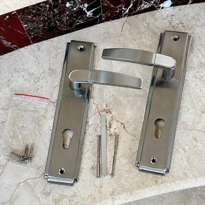 May include: A set of two rectangular brushed stainless steel door handles with matching backplates, each featuring a keyhole cutout. The set includes a small plastic bag containing three metal screws and a central square spindle bar for installation.