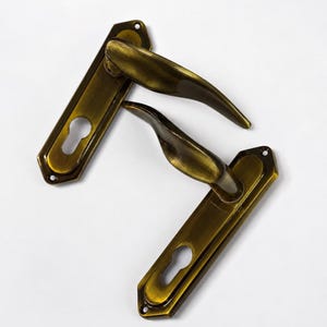Puede incluir: Dos manijas de puerta a juego con acabado en latón antiguo. Cada manija presenta una palanca curva unida a una placa rectangular con un corte para cerradura. El metal tiene una textura cepillada y oscura con orificios para tornillos.