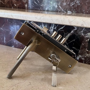 Op de afbeelding: Een messingkleurig metalen deurslotmechanisme met een deurkruk, een rechthoekige voorplaat en een cilinderslot met een ingestoken sleutel. Een ring met diverse zilverkleurige sleutels hangt aan de hoofdsleutel in de cilinder.