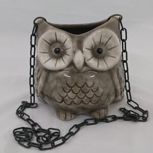 Op de afbeelding: Een grijze keramische uilvormige plantenbak met grote witte cirkelvormige ogen met donkere pupillen, een kleine snavel en gestructureerde veertjes. De plantenbak hangt aan een donkere metalen ketting bevestigd aan twee lussen aan de zijkanten.