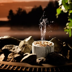 May include: A small, grey stone fountain with water spraying upwards, surrounded by dark rocks. The fountain is filled with small, brown pebbles. The background features a blurred landscape with a dark forest and a dusky sky.