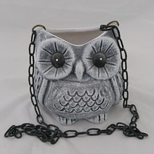 May include: A gray and white owl-shaped hanging planter with a textured finish. The planter features large dark eyes with white pupils and a carved feather pattern on the body. It is suspended by two black metal chains attached to brass-colored rings.