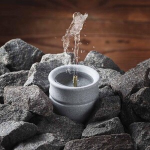 May include: A small, gray stone fountain with water spraying upwards. The fountain is surrounded by gray rocks, with a wooden background. The water stream is clear and the fountain has a brass nozzle.