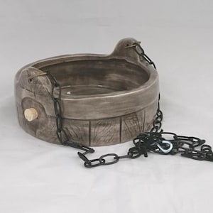 May include: A round, gray ceramic hanging planter with a textured wood-grain pattern on the exterior. The planter features a small, light-colored circular plug on the side and is suspended by black metal chains with a silver-colored hook attachment.