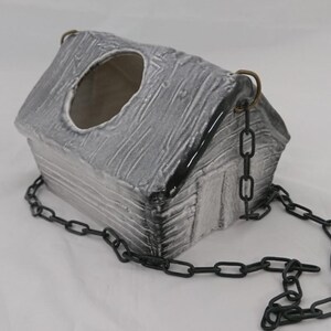 Puede incluir: Un recipiente de cerámica con forma de casa y acabado texturizado en gris y blanco desgastado. La estructura presenta una gran abertura circular en el techo y está suspendida por cadenas de metal negro unidas a anillos de color bronce.