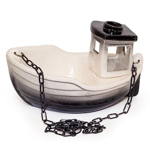 Peut inclure: Un bol en céramique en forme de bateau avec un intérieur crème et un extérieur dégradé noir et blanc. L'objet présente une cabine surélevée et une chaîne en métal noir fixée à la proue et à la poupe, retombant sur le devant du récipient.