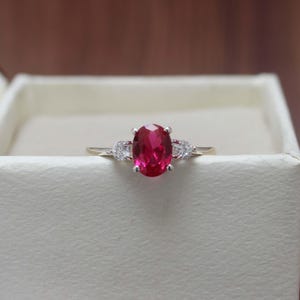May include: A silver-toned ring featuring a central oval-cut vibrant red gemstone held in a four-prong setting, flanked by two smaller clear round-cut stones on each side, set on a thin polished metal band resting inside a textured white box.