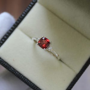 Könnte beinhalten: Ein silberner Ring mit einem großen, quadratisch geschliffenen, tiefroten Edelstein als Mittelpunkt. Zwei kleinere, klare Edelsteine flankieren den Hauptstein. Der Ring liegt in einer schwarzen Schmuckschatulle mit cremefarbenem Innenraum.
