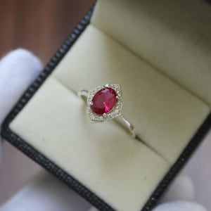 Puede incluir: Anillo de plata con una gran gema de rubí ovalada, rodeada de pequeños diamantes blancos. El anillo tiene un engaste decorativo de inspiración floral y se muestra en una caja negra.
