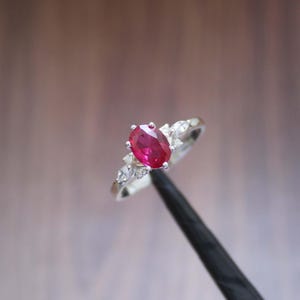 Może przedstawiać: Srebrny pierścionek z dużym, owalnym rubinem w kolorze intensywnej czerwieni. Mniejsze, przezroczyste kamienie szlachetne osadzone są po obu stronach rubinu, dodając blasku. Pierścionek jest trzymany przez czarny przedmiot. Biżuteria.