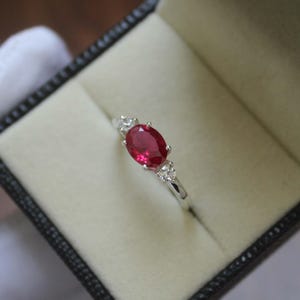 May include: A silver ring featuring a large oval ruby gemstone at the centre, flanked by two smaller clear stones. The ring is presented in a black jewellery box with a cream-coloured interior.