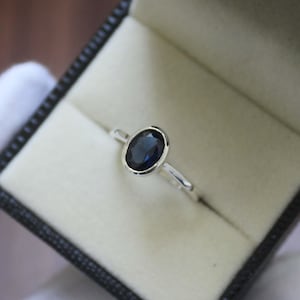 Könnte beinhalten: Ein silberner Ring mit einem dunkelblauen, ovalen Edelstein in einer Zargenfassung. Der Ring wird in einer schwarzen Schmuckschatulle mit cremefarbenem Innenraum präsentiert. Das Band des Rings ist schmal und schlicht.