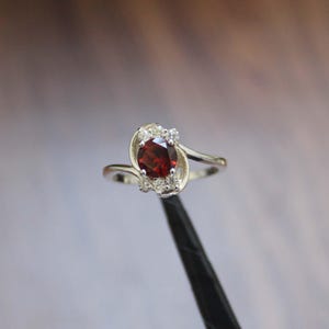 May include: A silver ring with a central, oval-cut, deep red gemstone, likely a garnet, encircled by smaller, clear stones. The ring's design is a swirling pattern with a polished finish, presenting a classic and elegant aesthetic.