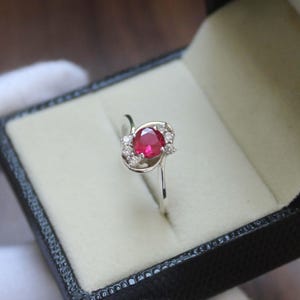 May include: A silver ring featuring a central, round, red gemstone, encircled by smaller, clear stones. The ring is presented in a black jewellery box with a light-coloured interior.