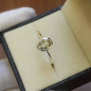 Pode incluir: Um anel de prata com uma pedra preciosa oval verde claro em uma moldura de bisel. O anel é exibido dentro de uma caixa de joias preta com interior creme. A pedra preciosa tem um tom verde pálido.