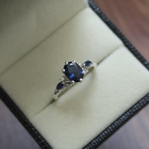 Puede incluir: Un anillo de plata con una gran piedra preciosa ovalada de color azul oscuro como pieza central. El anillo presenta piedras azules más pequeñas y piedras de acento transparentes en la banda, engastadas en un patrón decorativo. El anillo se muestra en una caja de joyería negra.